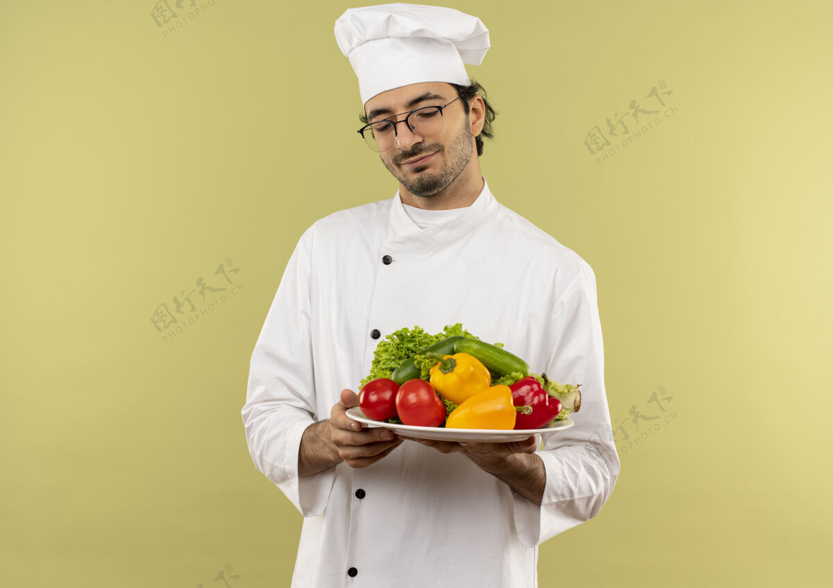 烹饪年轻的男厨师闭着眼睛 穿着厨师制服 戴着眼镜 把蔬菜放在隔离在绿色墙上的盘子里男性盘子厨师