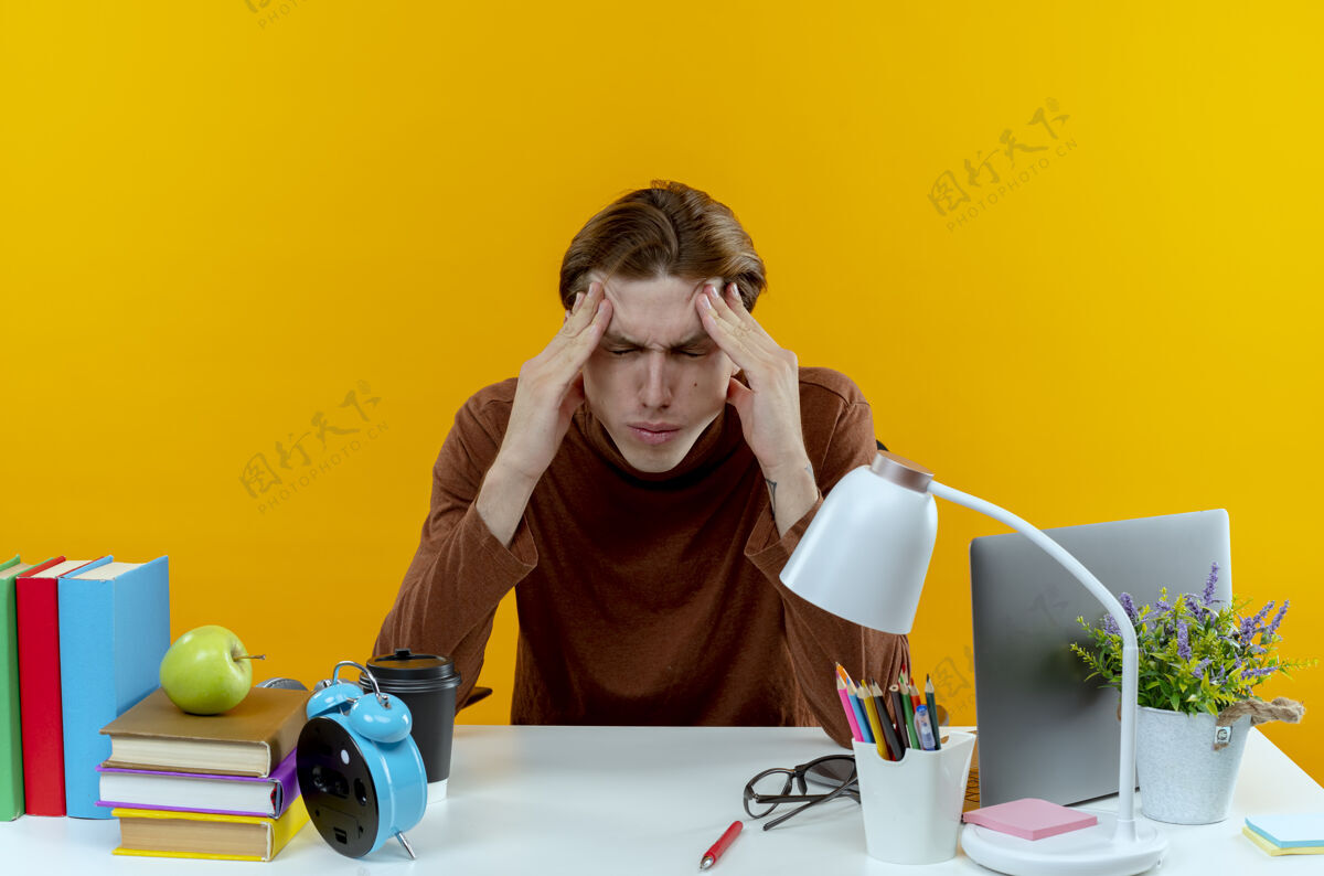 坐着一个疲惫的小男孩闭着眼睛坐在书桌旁 手里拿着学习工具 把眼睛孤立地放在黄色的墙上手男孩工具