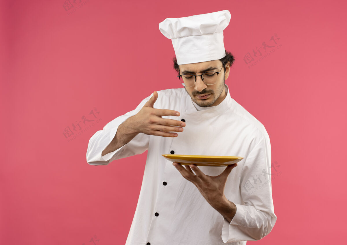 眼镜年轻的男厨师闭着眼睛 穿着厨师制服 戴着眼镜 假装嗅着隔离在粉红色墙上的食物食物嗅封闭