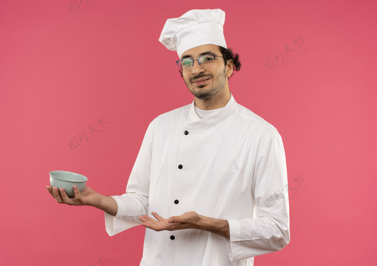 请高兴的年轻男厨师穿着厨师制服 拿着眼镜 用手指着隔离在粉红色墙上的碗男拿着碗