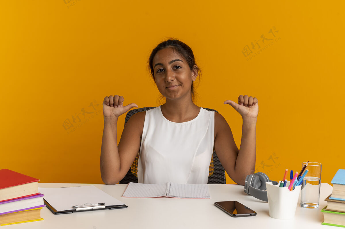 工具坐在书桌旁拿着学习工具的年轻女孩高兴地指着自己孤零零地站在橙色的墙上学校书桌她自己