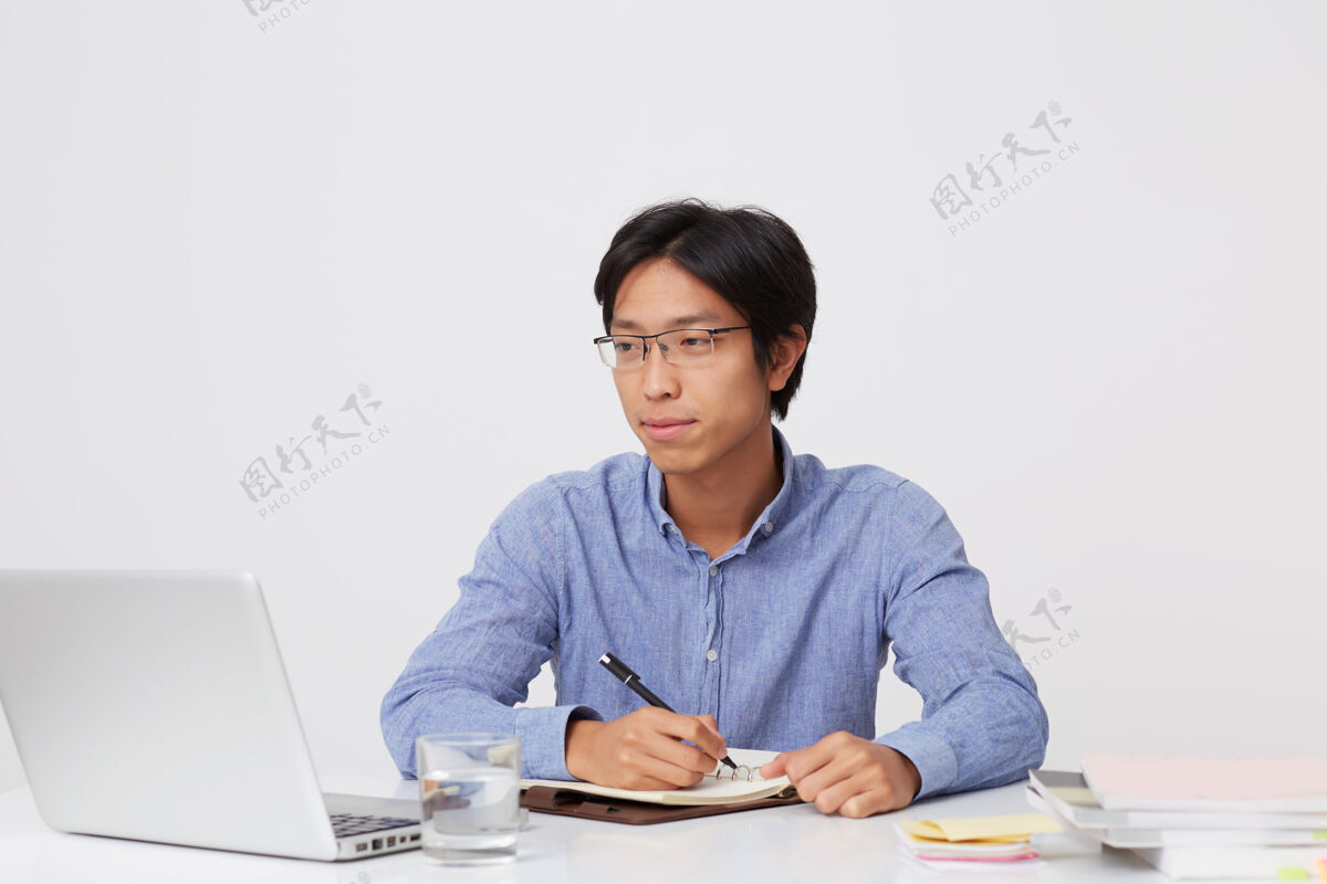 认真一位戴眼镜的亚洲年轻富商 面带沉思 在笔记本上写字 在桌旁工作 笔记本电脑盖在白墙上计划桌子亚洲人