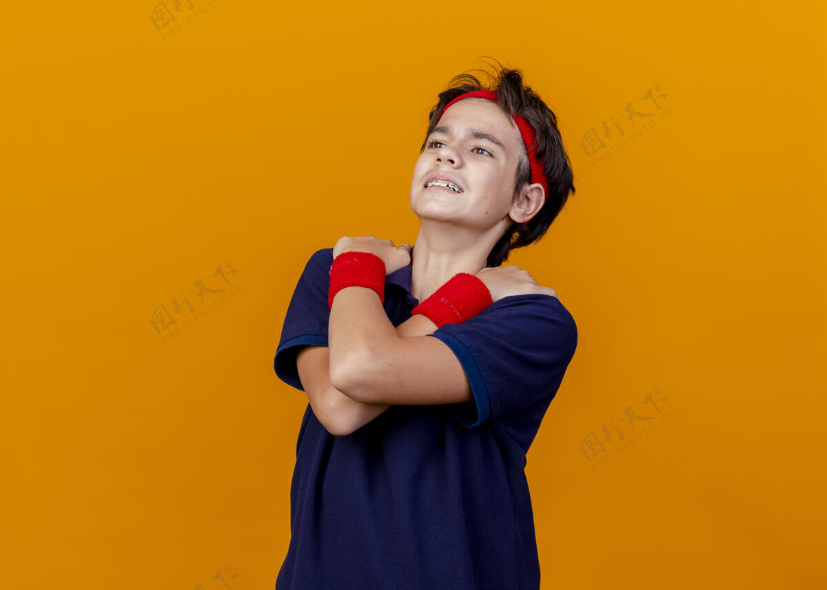 背带年轻帅气的运动男孩戴着头带和护腕 戴着牙套 看起来笔直 双手交叉放在肩膀上 橙色背景上 留有复制空间手男孩运动