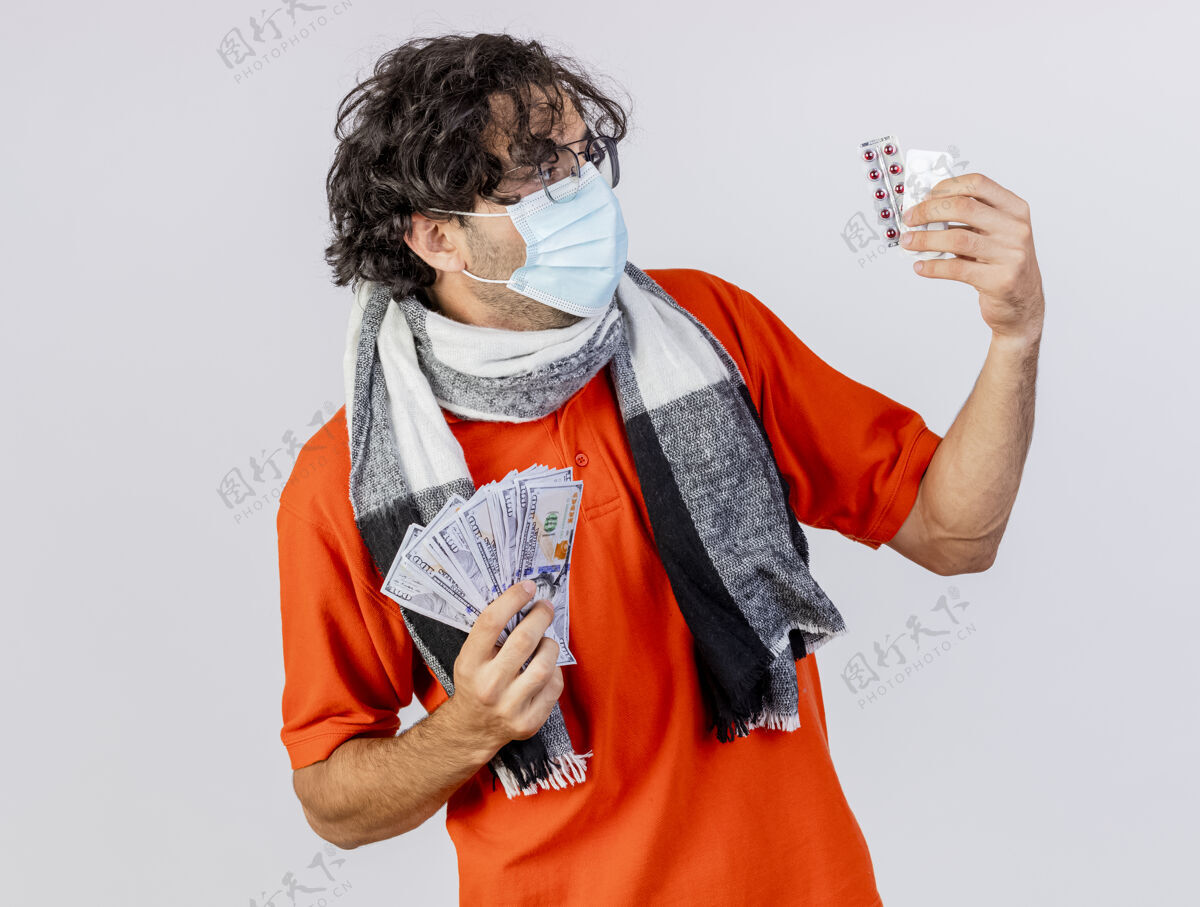 年轻年轻的白人病人戴着眼镜 围巾和口罩 手里拿着钱和药丸 看着隔离在白色背景上的药丸面具医疗疾病