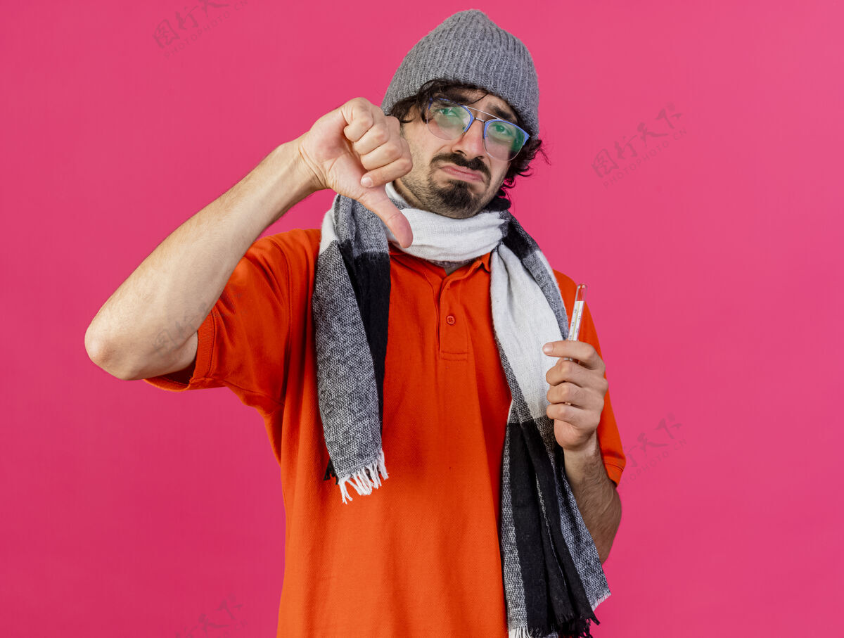 温度计年轻的白种人 戴着眼镜 戴着帽子 戴着围巾 拿着温度计 看着相机 拇指朝下 在深红色的背景下 留着复印空间放下围巾不愉快