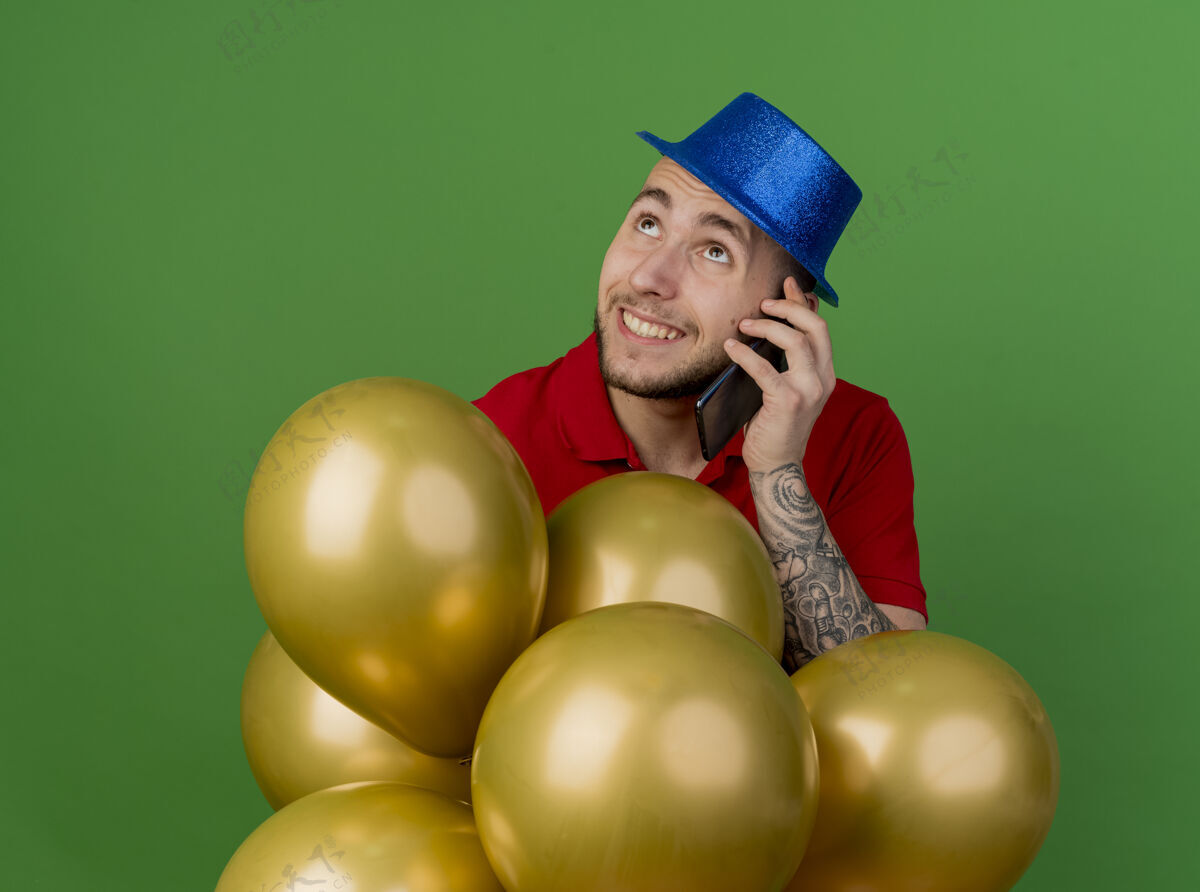 站着微笑着的年轻英俊的斯拉夫党人戴着党的帽子站在气球后面看着电话交谈绿色背景隔离帽子微笑气球