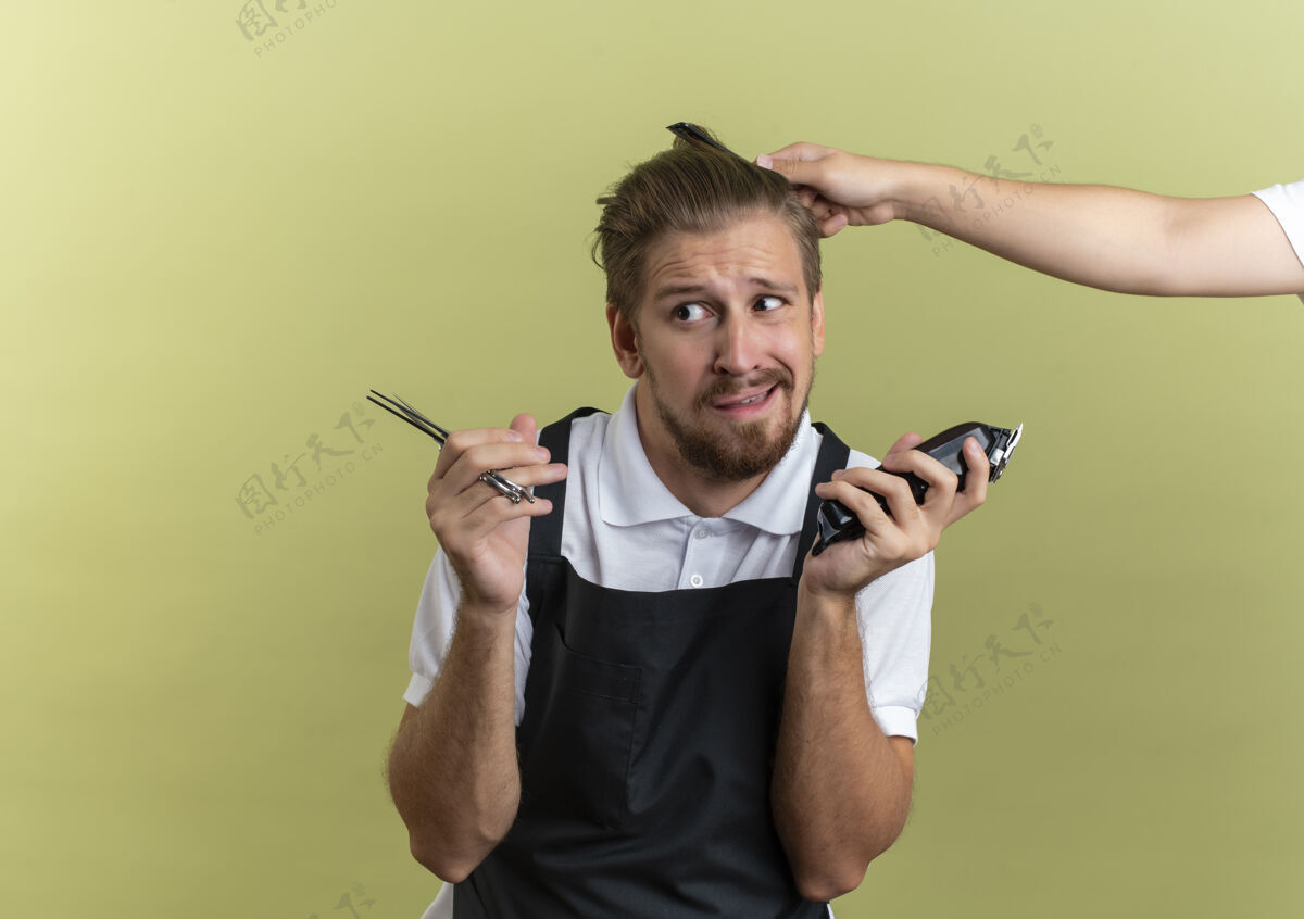 剪刀困惑的年轻英俊的理发师拿着剪刀和剪子 看着旁边有人梳着他的头发 橄榄绿的头发上孤立着复制空间困惑有人理发师