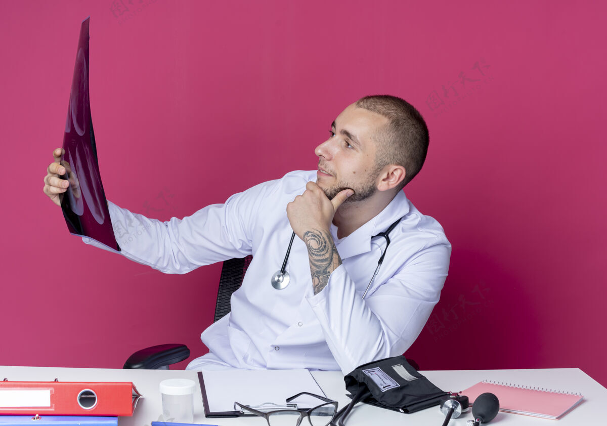 下巴自信的年轻男性医生穿着医用长袍和听诊器坐在办公桌旁 手里拿着工作工具 手放在粉红色的下巴上看x光片坐着穿着X光