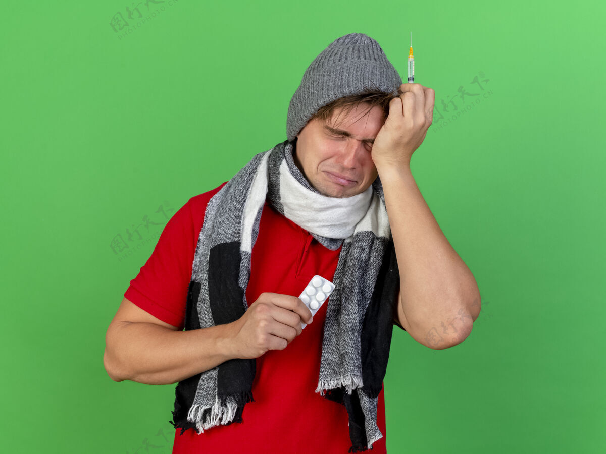 金发悲伤又害怕的年轻英俊的金发病患戴着冬天的帽子和围巾拿着一包药片和注射器触摸着头闭着眼睛隔离在绿色的背景和复制空间年轻悲伤围巾