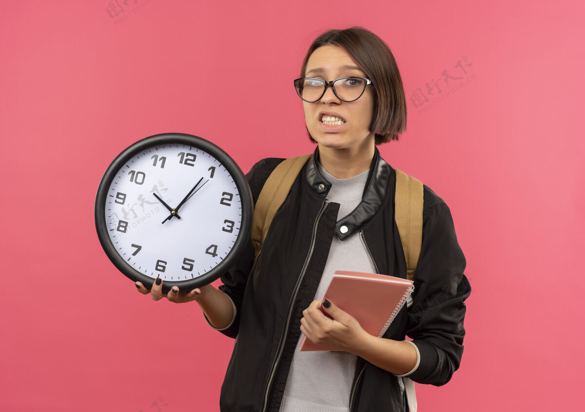 粉色焦急的年轻女学生戴着眼镜 背着手袋拿着时钟和笔记本 孤立在粉红色的纸上纸条急女孩