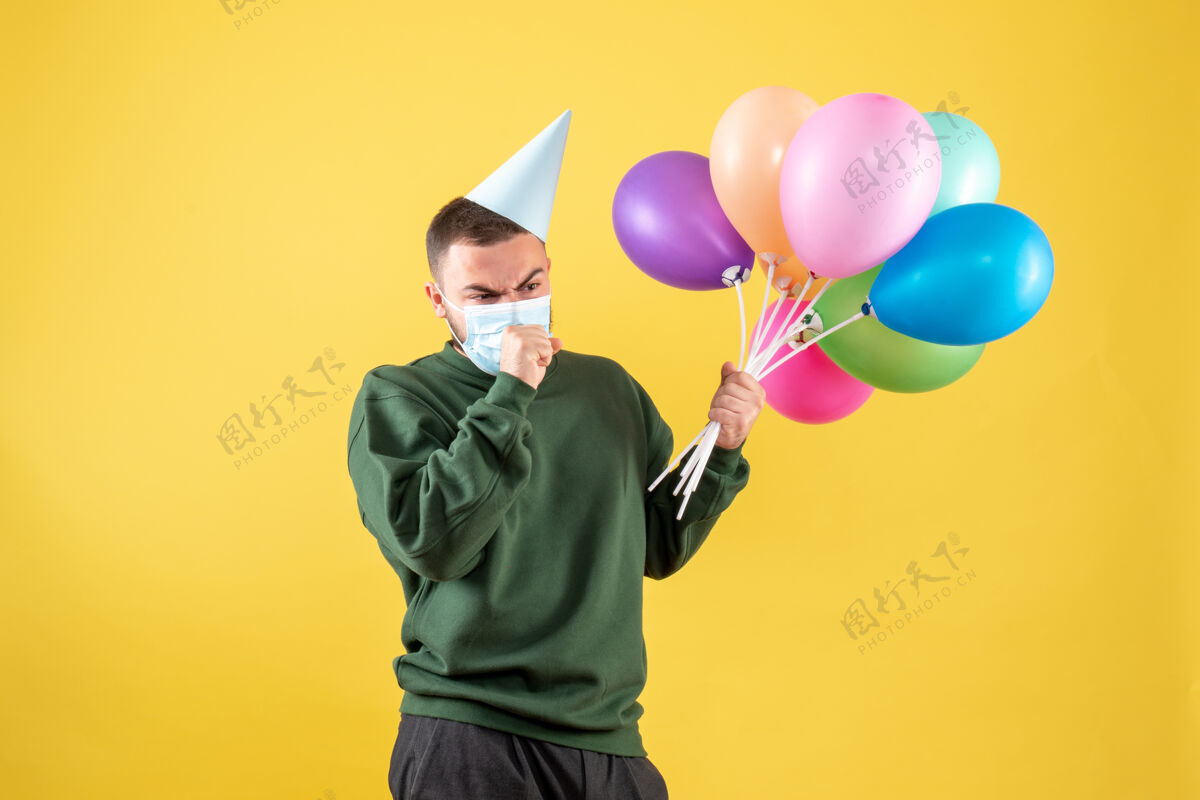 乐趣正面图黄色背景下 戴着面具手持彩色气球的年轻男性举行年轻男性人