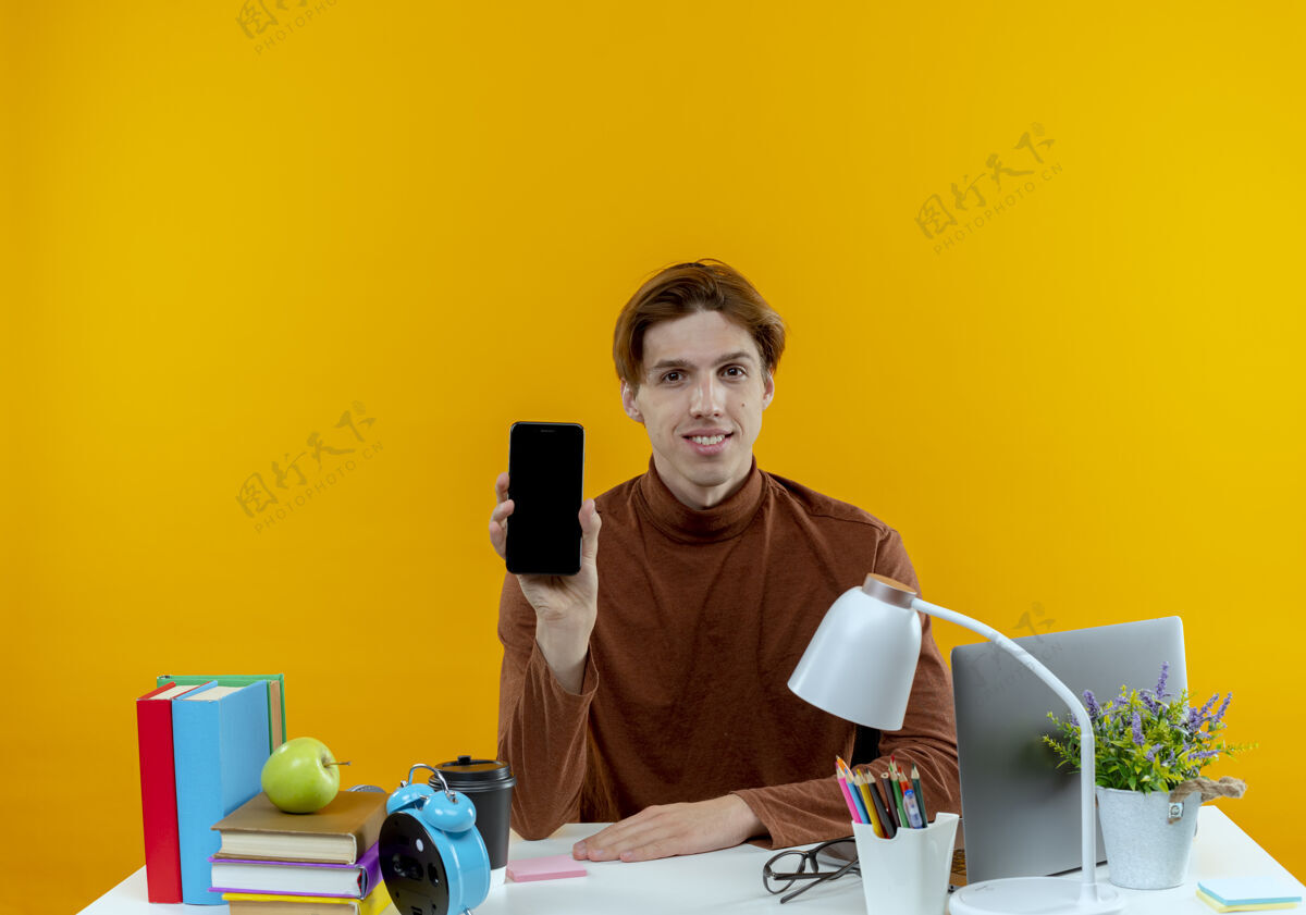 抱着高兴的年轻学生男孩坐在书桌旁 拿着学习工具 把电话孤立在黄色的墙上坐着学生工具