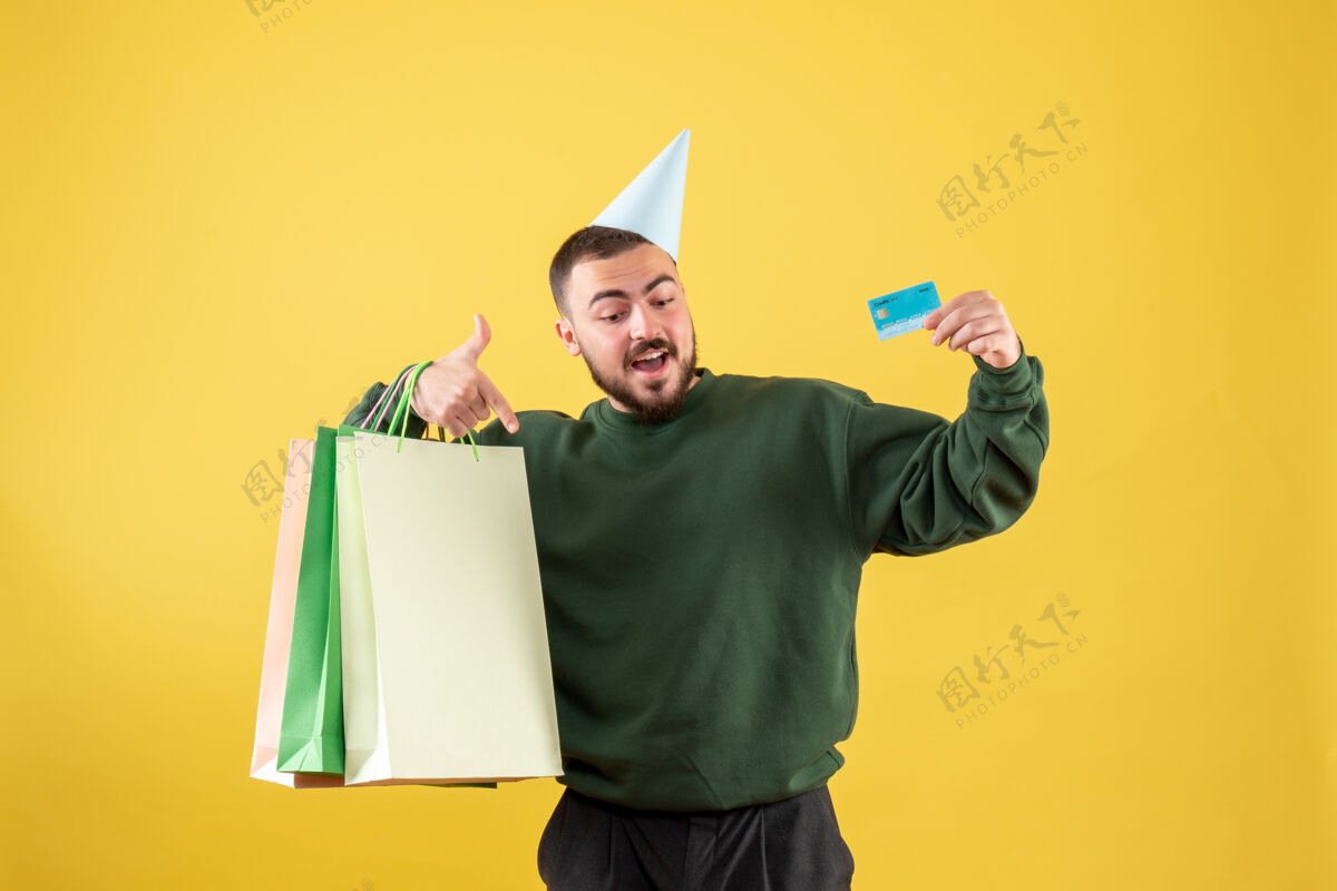 购物正面图年轻男性手持黄色背景的银行卡和购物包公园圣诞节聚会