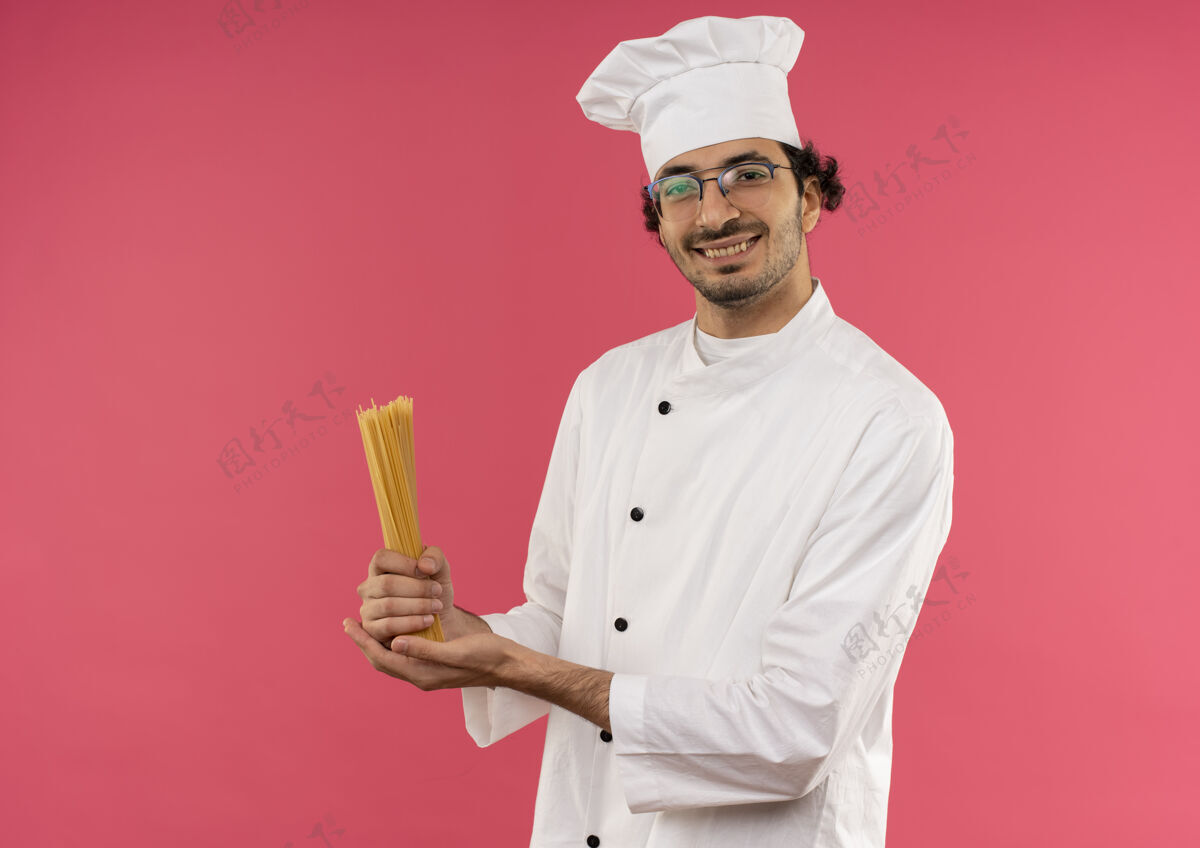 眼镜面带微笑的年轻男厨师穿着厨师制服 戴着眼镜 把意大利面孤立地放在粉红色的墙上男年轻意大利面