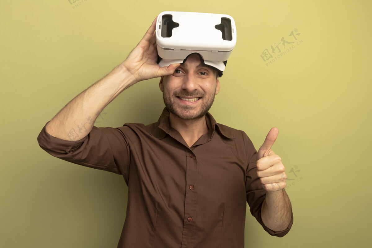 相机微笑的白人年轻人头戴虚拟现实耳机抓着它看相机显示拇指向上橄榄绿背景与复制空间隔离抓住橄榄耳机