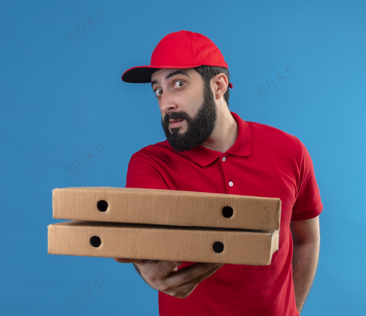 制服印象深刻的年轻英俊的白人送货员穿着红色制服和帽子伸出比萨饼盒对着镜头隔离在蓝色送货穿着男人