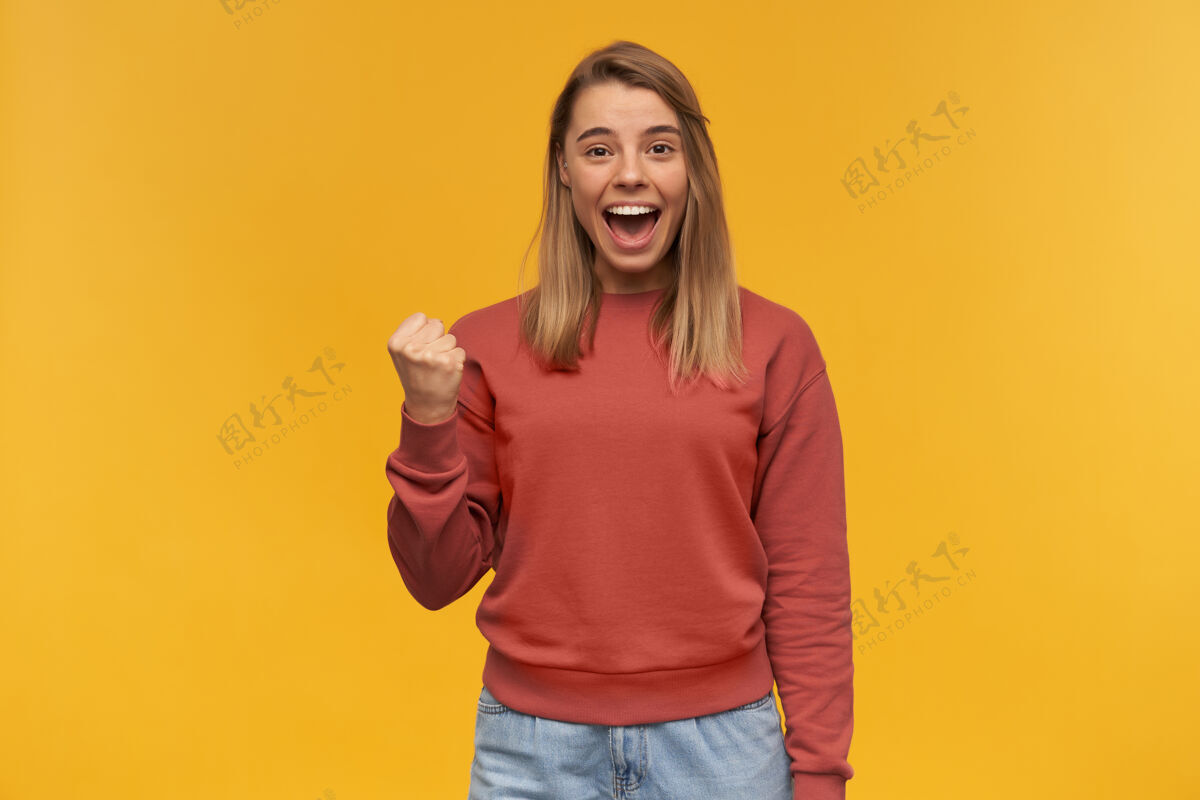 运动衫穿着休闲服的快乐成功的年轻女子在黄色的墙壁上展示赢家的姿态和喊叫情绪女性微笑