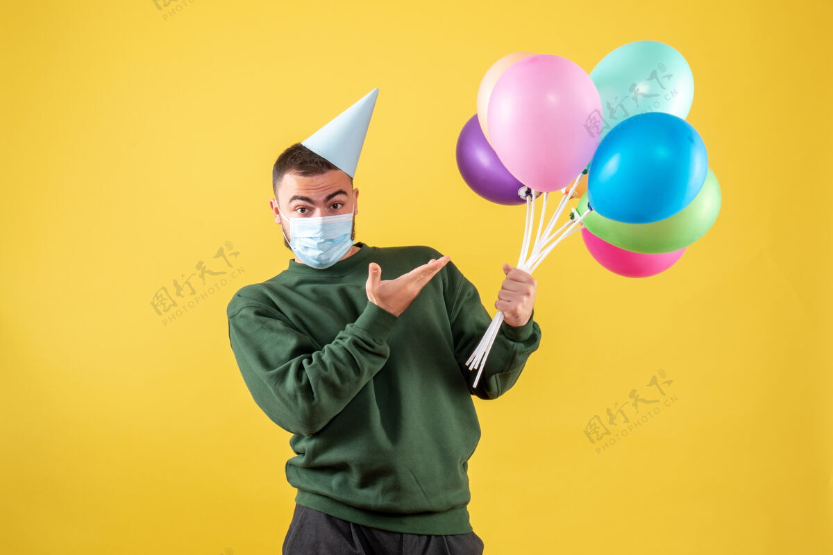 病毒正面图：黄色背景上戴着面具手持彩色气球的年轻男性庆祝人面具