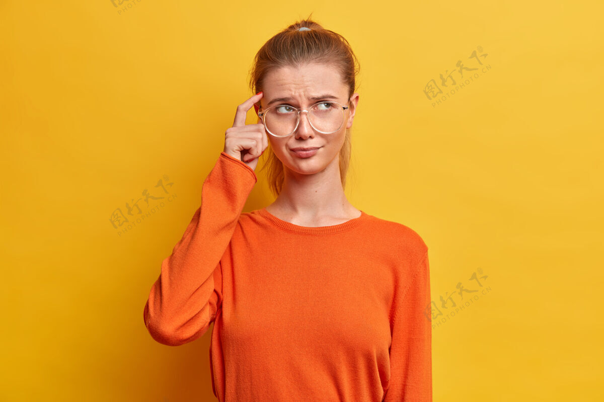 模特体贴严肃的女人的肖像手指放在太阳穴上 试图专注于某事 穿着休闲的橙色套头衫 摆姿势女人眼镜女性