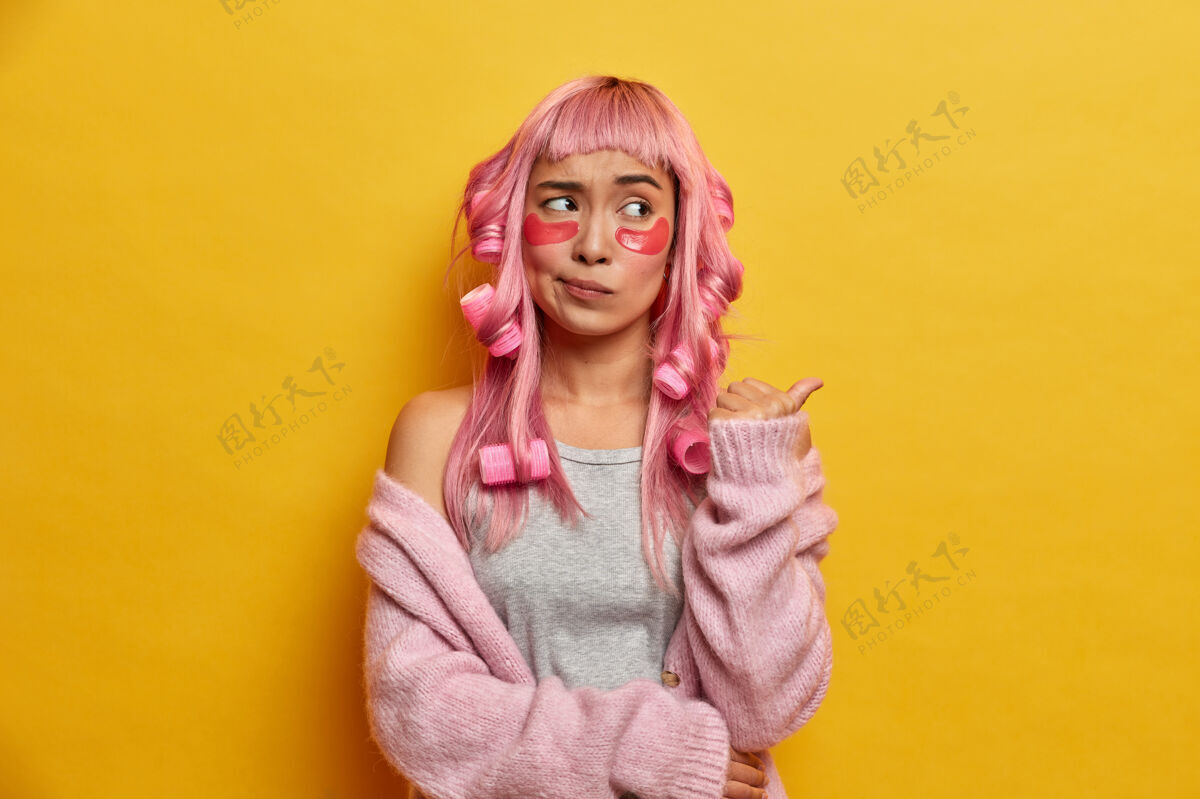 严肃迷惑不解的亚洲女孩的室内镜头有着粉红色的长发 用卷发器做卷发 指着复制空间 有不悦的表情室内粉色垫子