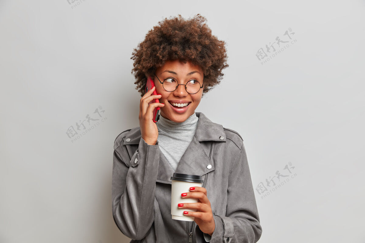 女士快乐无忧无虑的年轻女子有着非洲式的发型 喜欢活泼的电话交谈 目光移开 端着一次性咖啡杯 戴着圆眼镜和灰色夹克姿势女士灰色