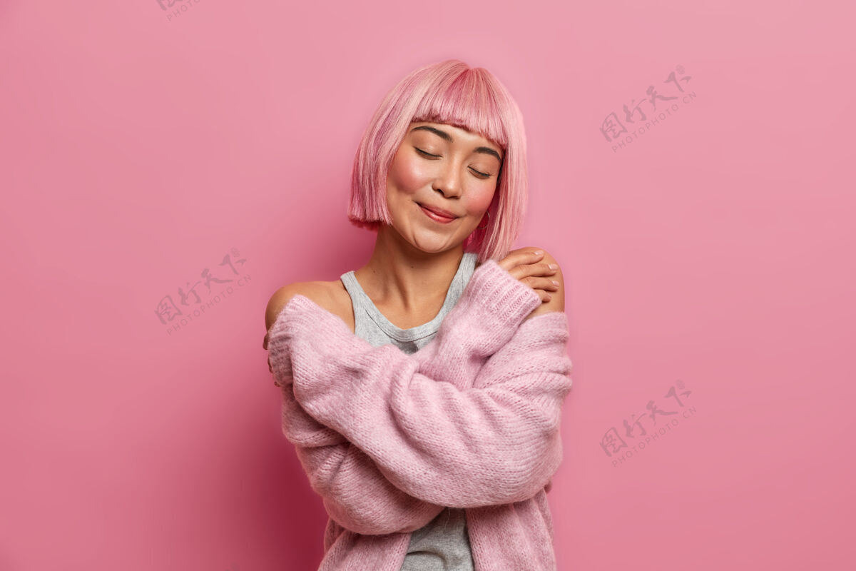 套头衫迷人漂亮的亚洲女人的特写肖像有粉红色的发型 拥抱自己 闭着眼睛站着 穿着暖和柔软的毛衣 站在室内梦想微笑舒适