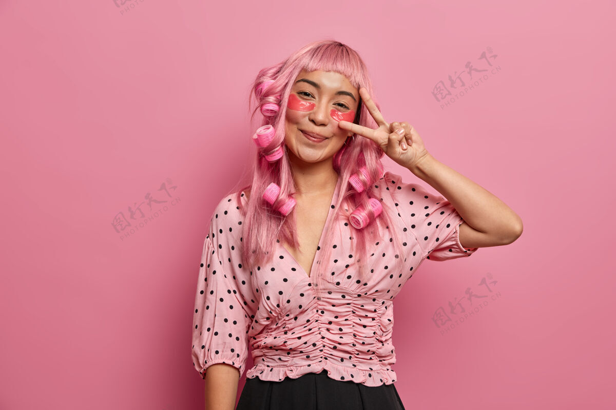 面部快乐的粉红色头发的女孩用眼罩来减少黑眼圈 使和平的迹象 戴卷发器有完美的卷发肖像美容圆圈