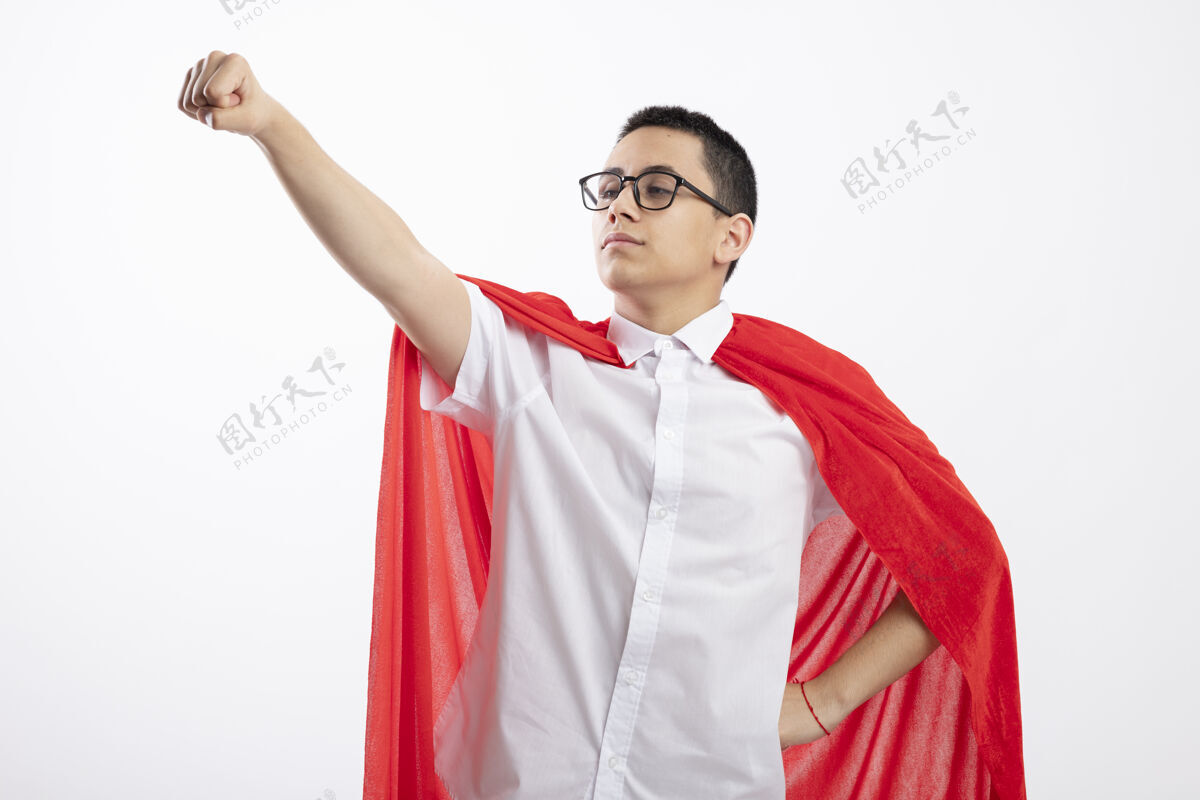 腰身穿红色斗篷 戴着眼镜 自信的年轻超级英雄男孩 手放在腰上 看着在白色背景下伸出的拳头男孩红色壁板