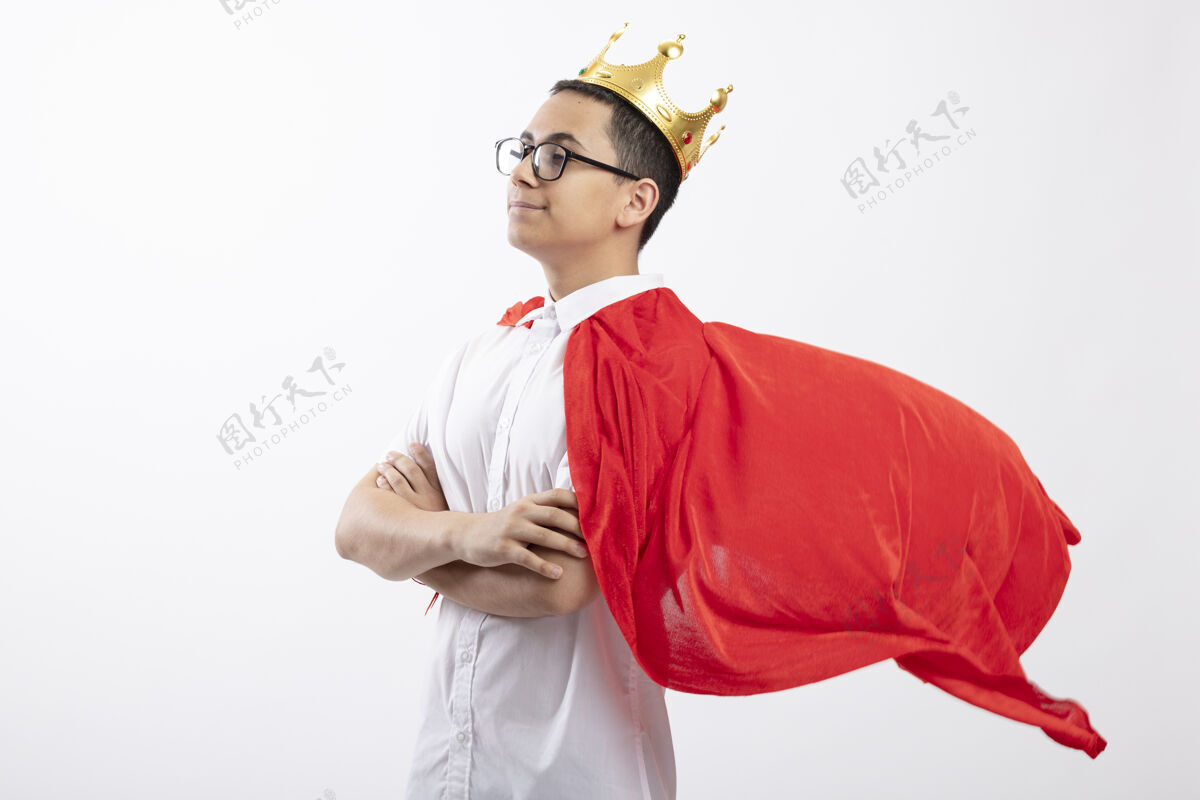 超级英雄自信的年轻超级英雄男孩穿着红色斗篷 戴着眼镜和皇冠 站在侧面视图中 以封闭的姿势 在白色背景下 看着笔直的孤立 有复制空间立场眼镜皇冠