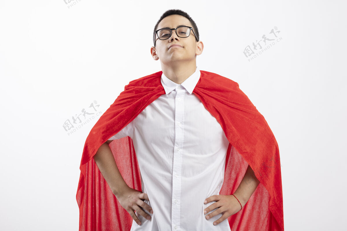 腰穿着红斗篷的自信的年轻超级英雄男孩戴着眼镜看着相机 手放在白色背景下的腰部手斗篷穿