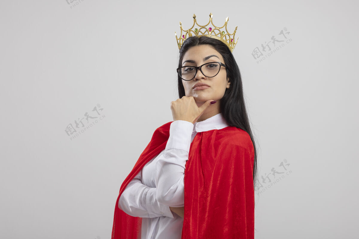 触摸可疑的年轻白种人超级英雄女孩戴着眼镜和皇冠站在侧视图看着相机触摸下巴隔离在白色背景与复制空间女孩下巴查看
