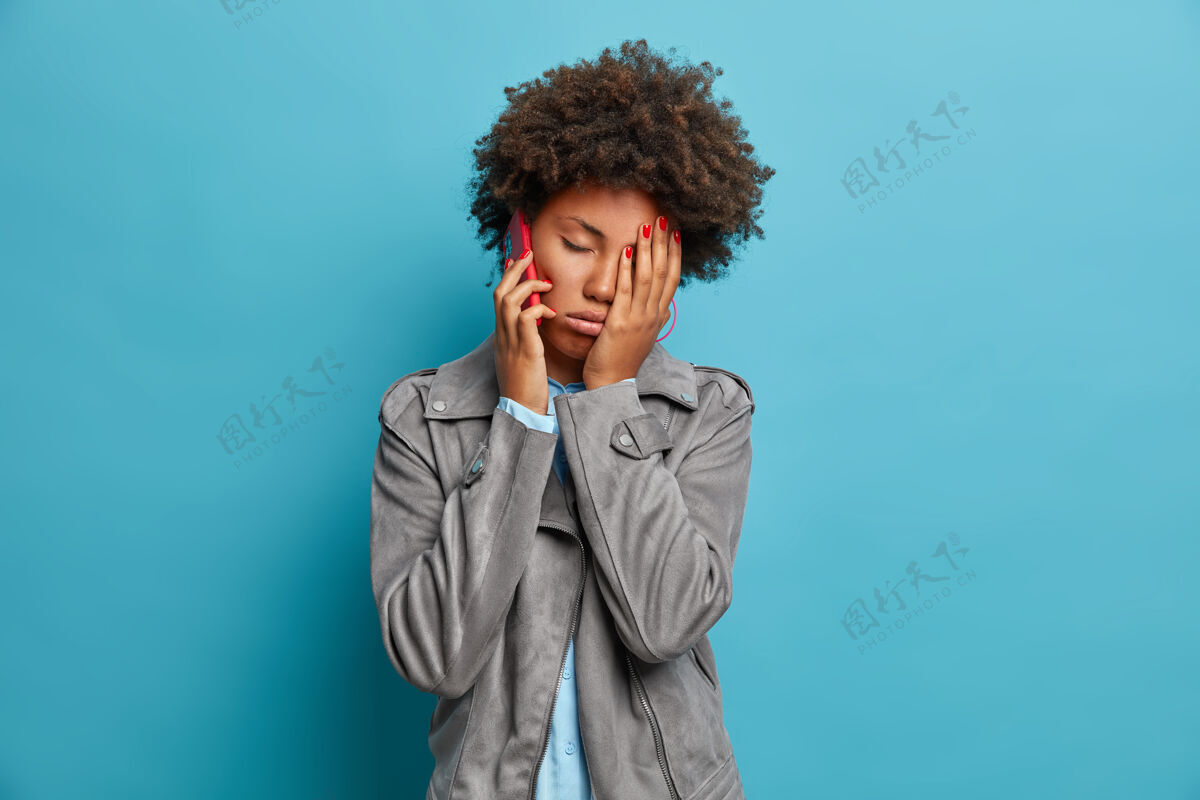 设备非洲头发的疲倦困倦的女人做脸手掌 感到疲倦和疲劳 有无聊的电话交谈 打电话给朋友 穿灰色夹克 摆姿势人抱着非洲