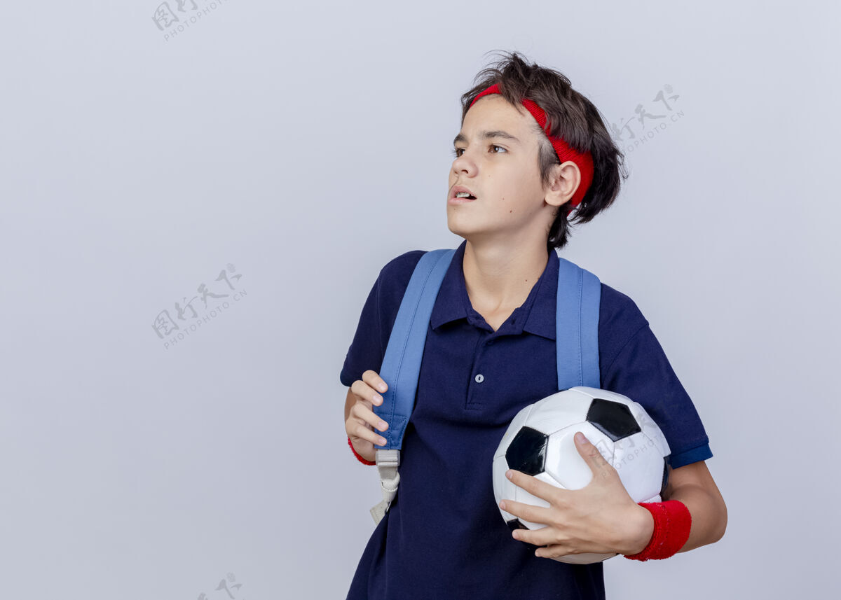 壁板自信的年轻帅气的运动男孩戴着头带和腕带 背上包着牙套 拿着包和足球的带子 看着在白色背景上有复制空间的隔离边包复制头带