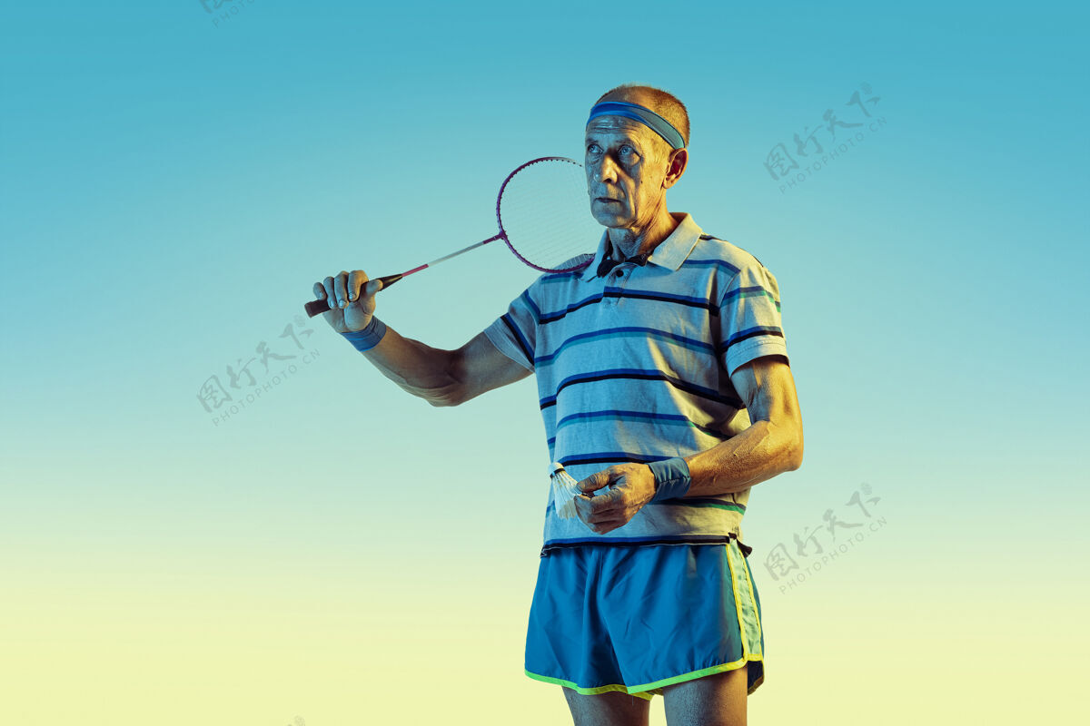 健康穿运动服的老人在梯度背景下打羽毛球 霓虹灯玩球拍运动