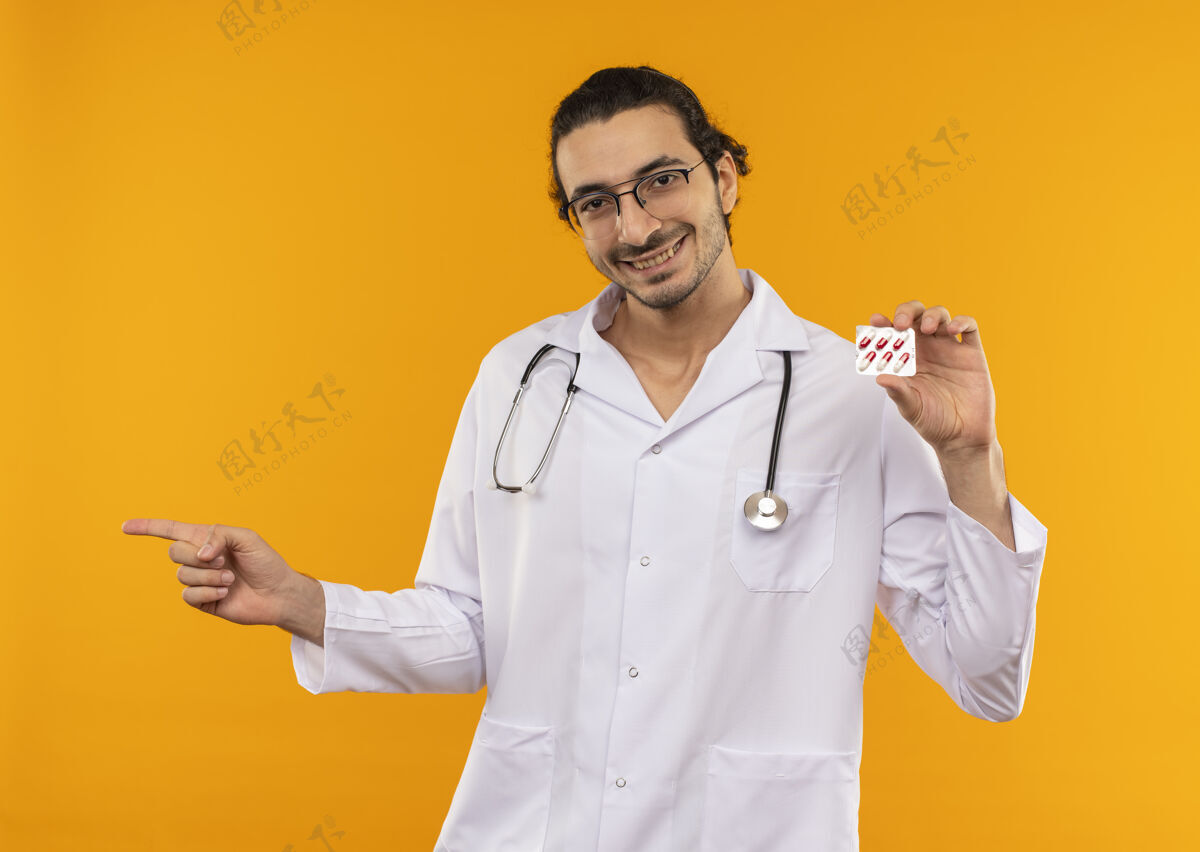 眼镜面带微笑的年轻医生戴着医用眼镜 穿着医用长袍 手持听诊器 指着黄色的一边年轻持有微笑