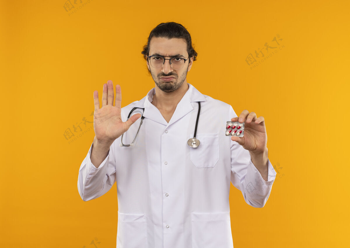 黄色戴着医用眼镜的年轻医生穿着带听诊器的医用长袍拿着药片 在黄色上显示停止手势医学药丸听诊器