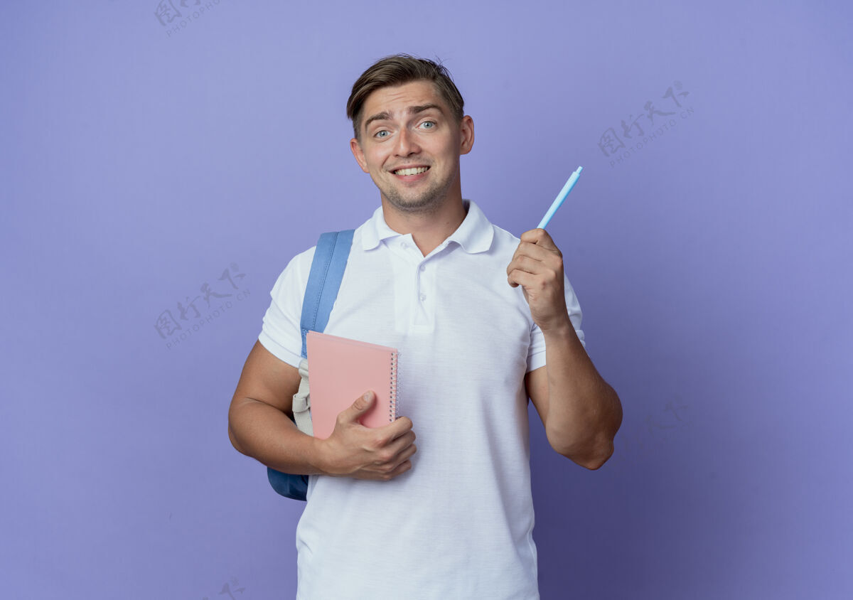 姿态印象深刻的年轻帅哥学生背着书包拿着笔记本 笔孤立在蓝色的墙上墙笔笔记本