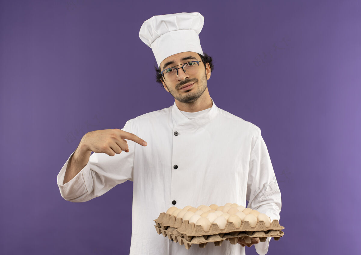 背景高兴的年轻男厨师穿着厨师制服 拿着一副眼镜 指着紫色的一批鸡蛋请厨师男性