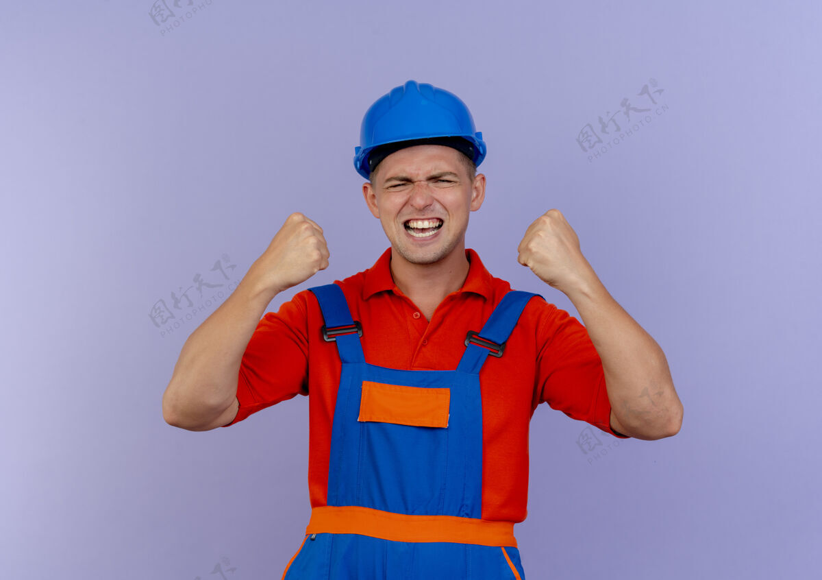 男性年轻的男建筑工人穿着制服 戴着安全帽 闭上眼睛 在紫色的地板上做着“是”的手势手势是的快乐
