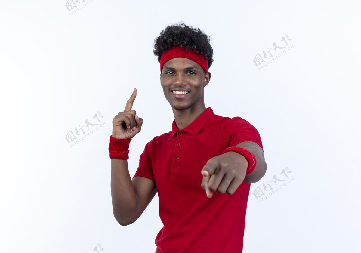 男人带着头带和腕带的年轻美籍黑人运动型男子微笑着向你展示手势 并指着白色的上方手势头带表情