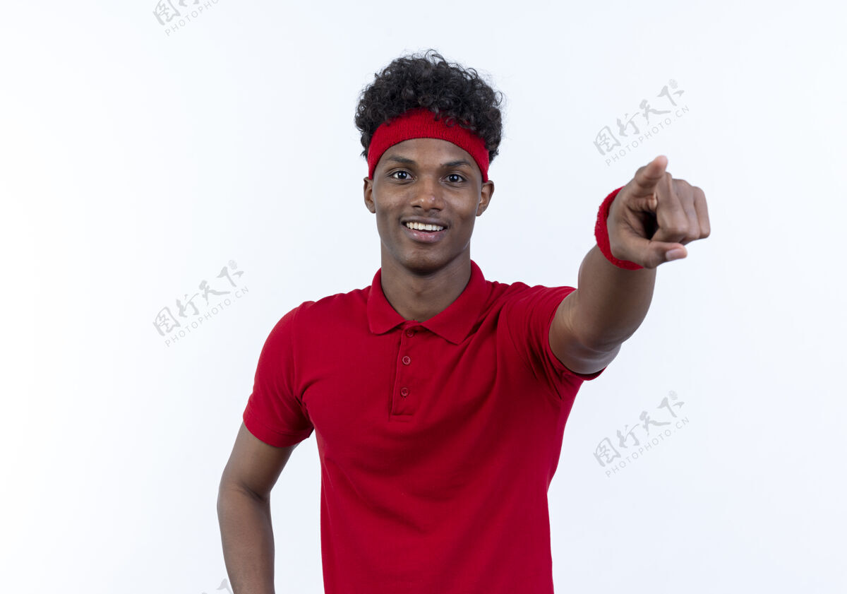 年轻带着头带和腕带的年轻黑人运动型男子微笑着指着隔离在白色背景下的摄像机头带相机腕带