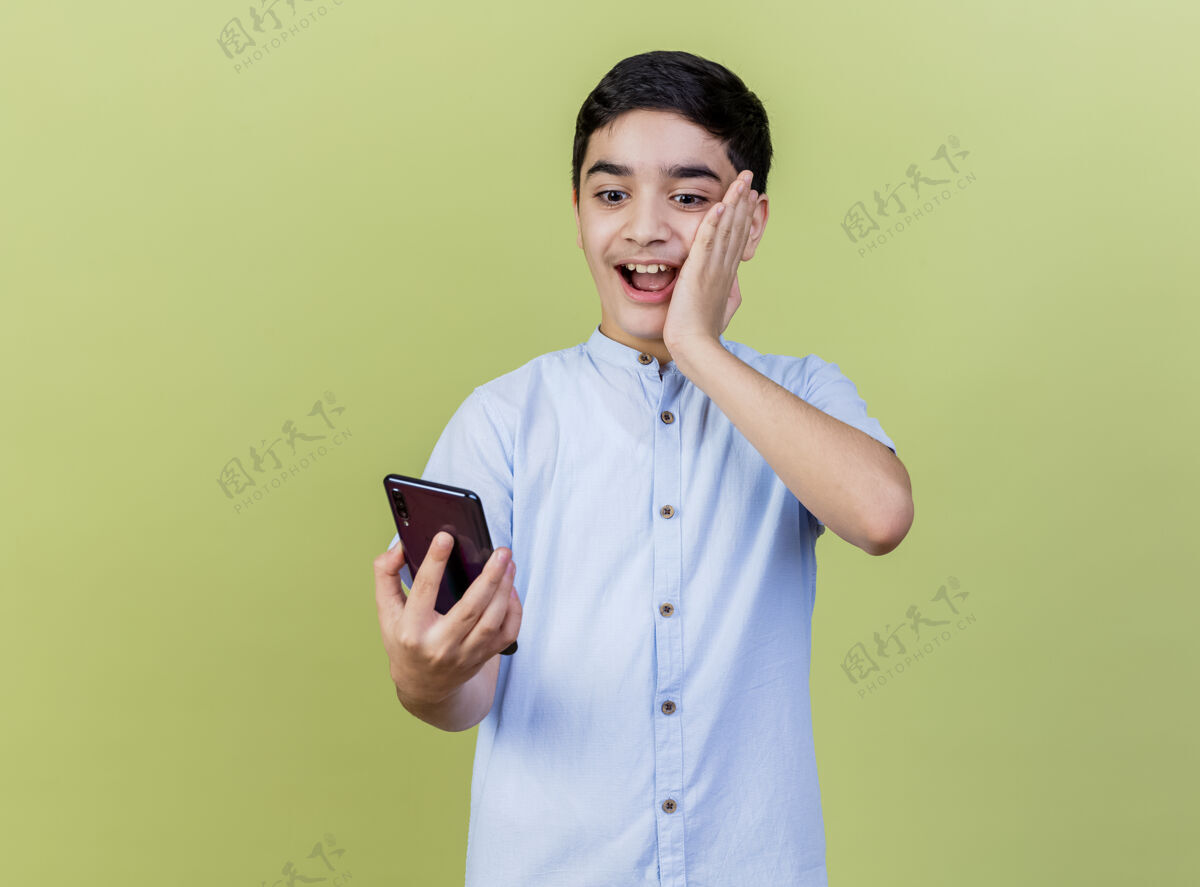 姿势印象深刻的小男孩拿着手机看着手放在脸上隔离在橄榄绿的墙上手电话表情