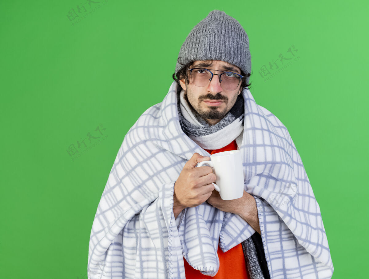 杯子虚弱的年轻病夫戴着眼镜 戴着冬天的帽子和围巾 裹着格子布 手里拿着一杯茶 抓着格子布 看着前面隔离在绿色墙上的墙人脸姿势