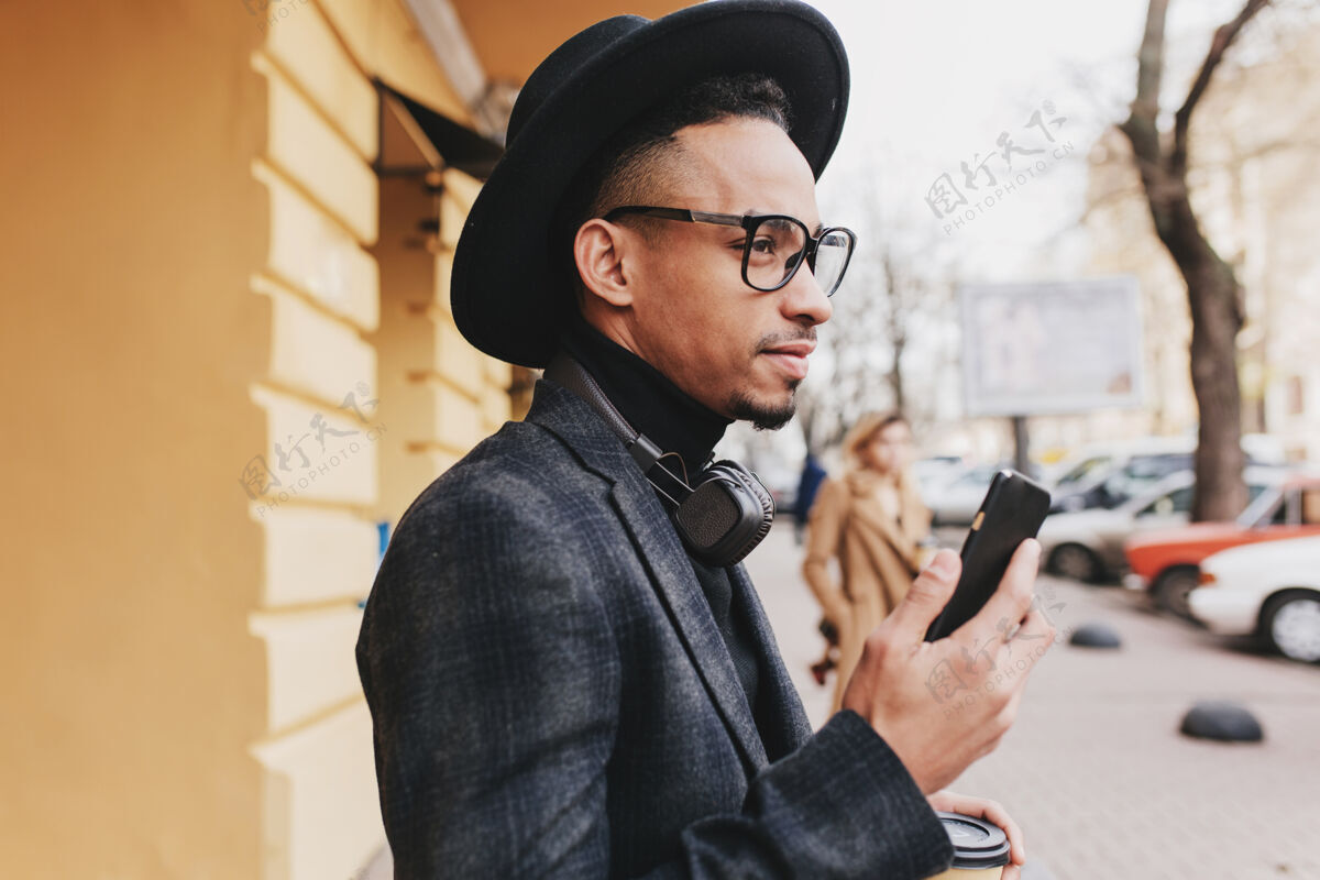 肖像一个严肃的棕色皮肤的男人拿着电话 看着街道一张穿着考究的非洲男人戴着帽子在咖啡馆附近等朋友的户外照片城镇男孩日光