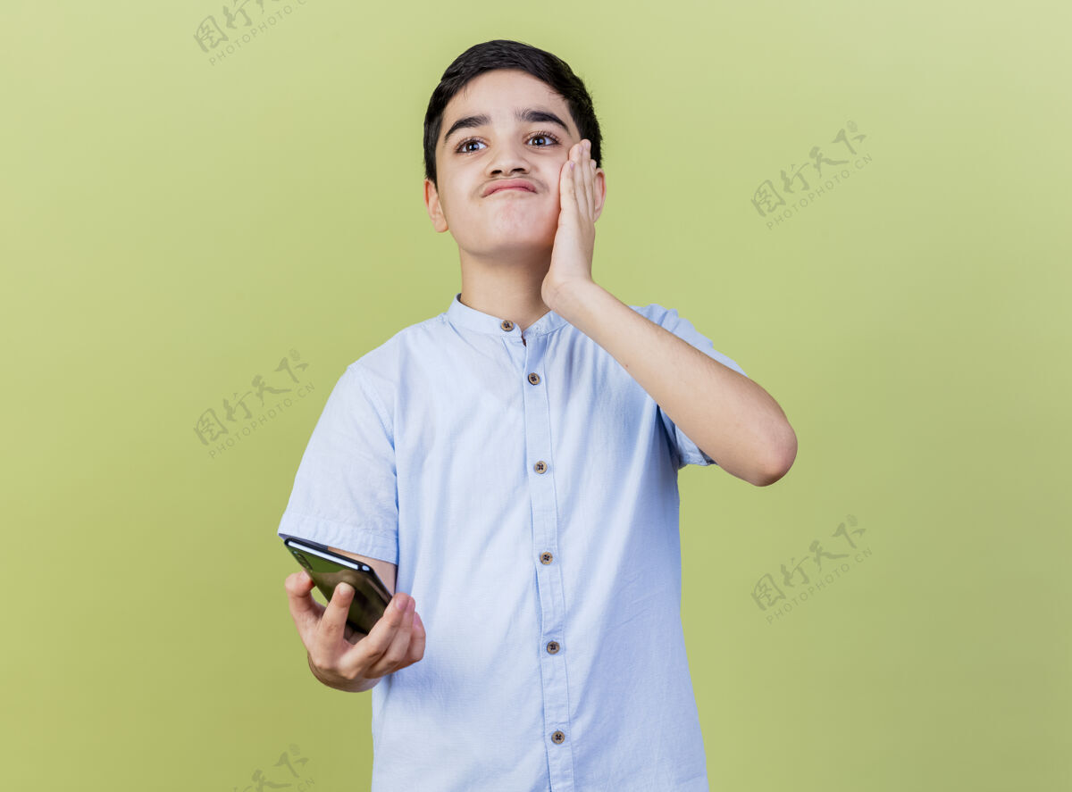 衣服未被释放的年轻男孩手持手机 手放在脸上看着隔离在橄榄绿墙上的一面市民年轻人