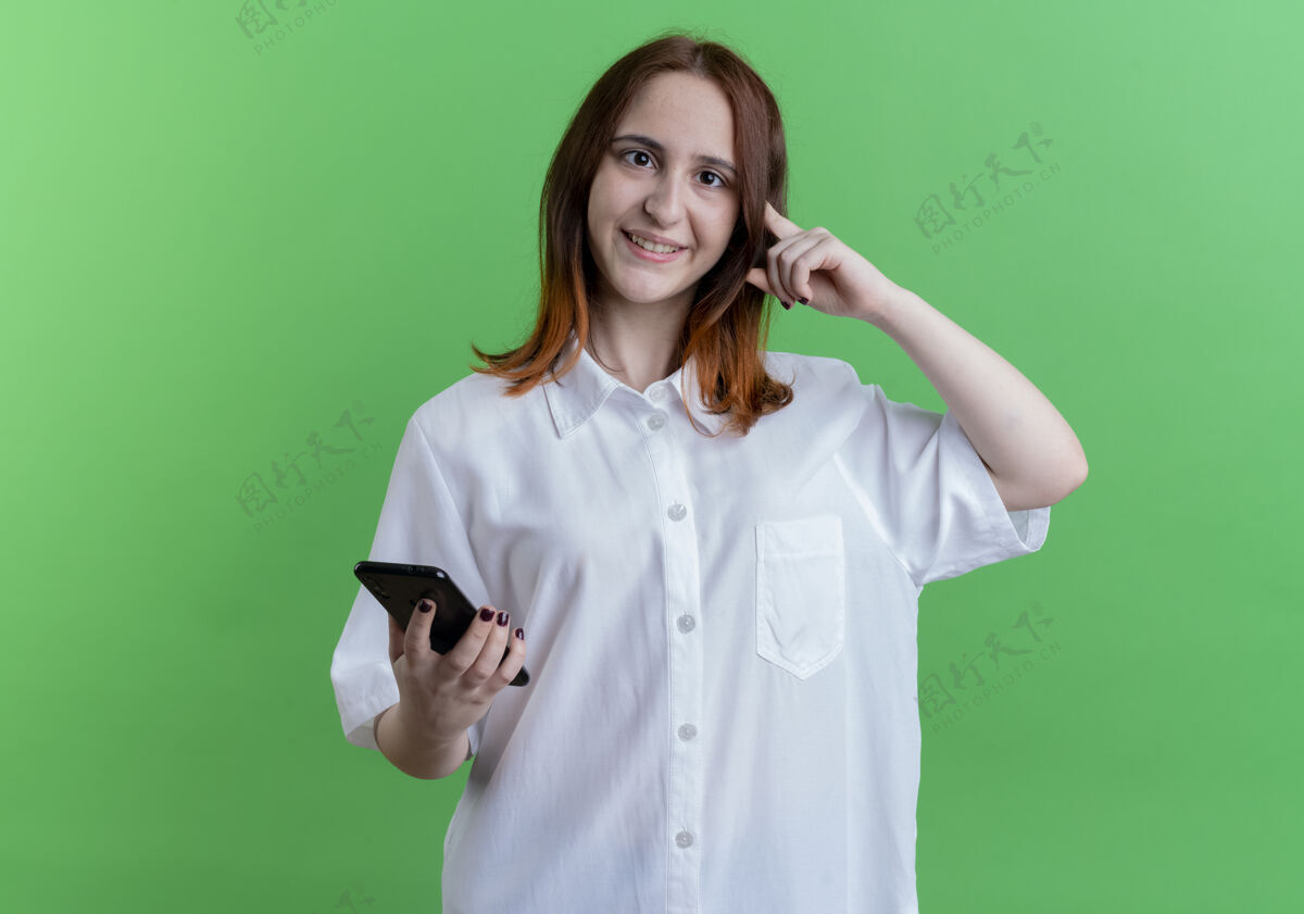 头微笑的年轻红发女孩拿着电话 把手指放在头上孤立的绿色女孩手指握着