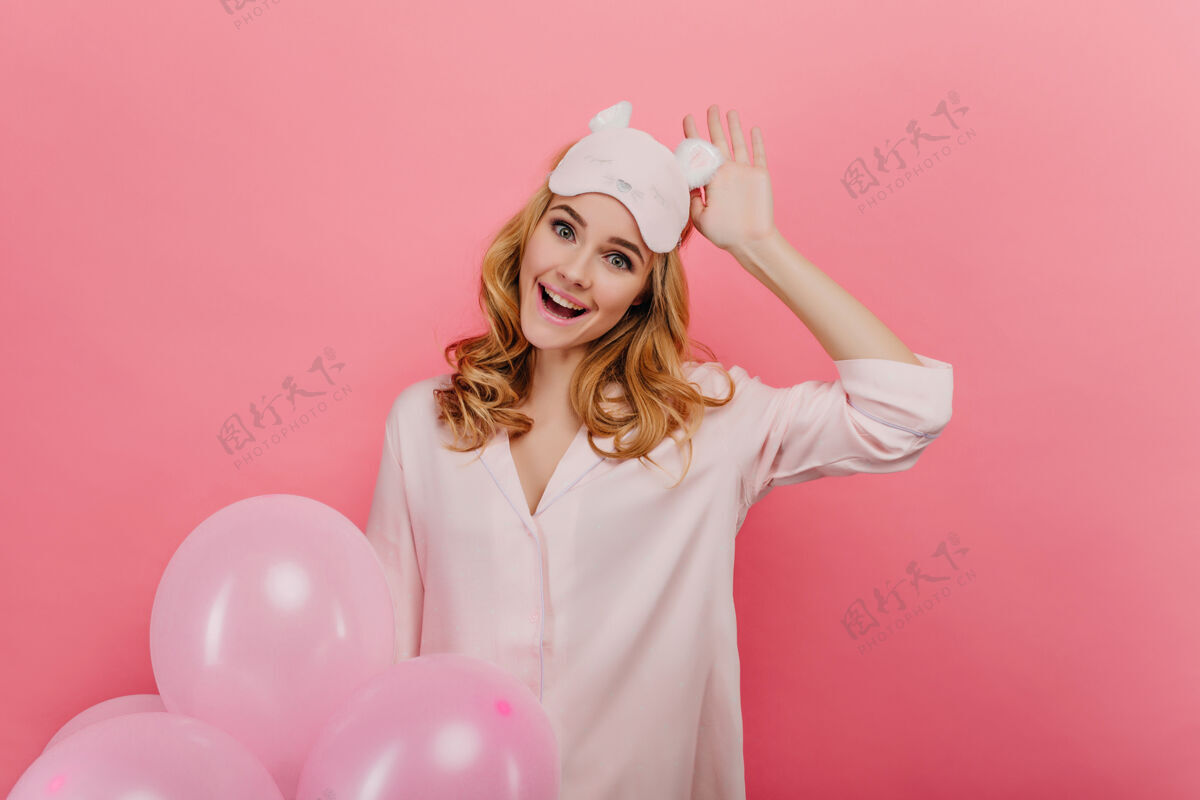 睡衣浪漫的欢笑女孩抚摸着她的睡眠面具 拿着明亮的气球美丽的卷发女士的室内肖像被隔离在粉红色的墙上氦气球气球肖像