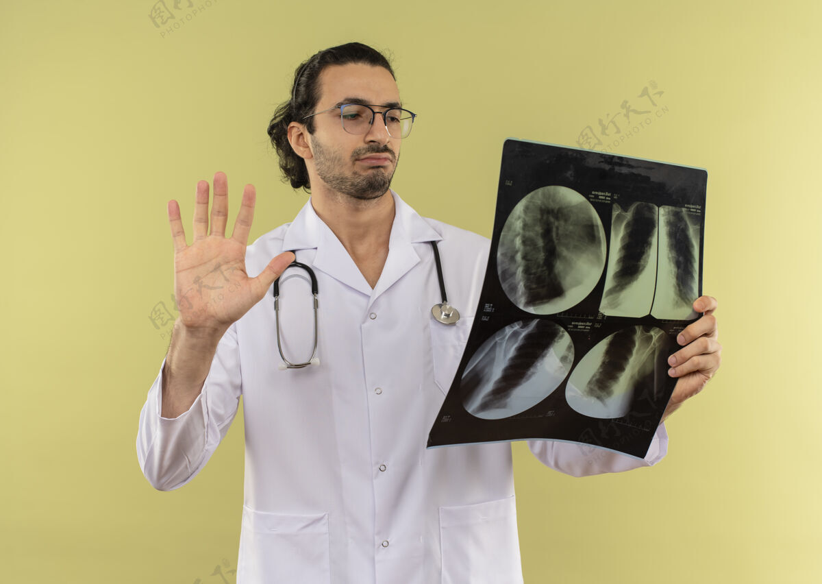 眼镜年轻的男医生戴着眼镜 穿着白色长袍 手持听诊器 看着x光片显示绿色的停止手势思考白色X光