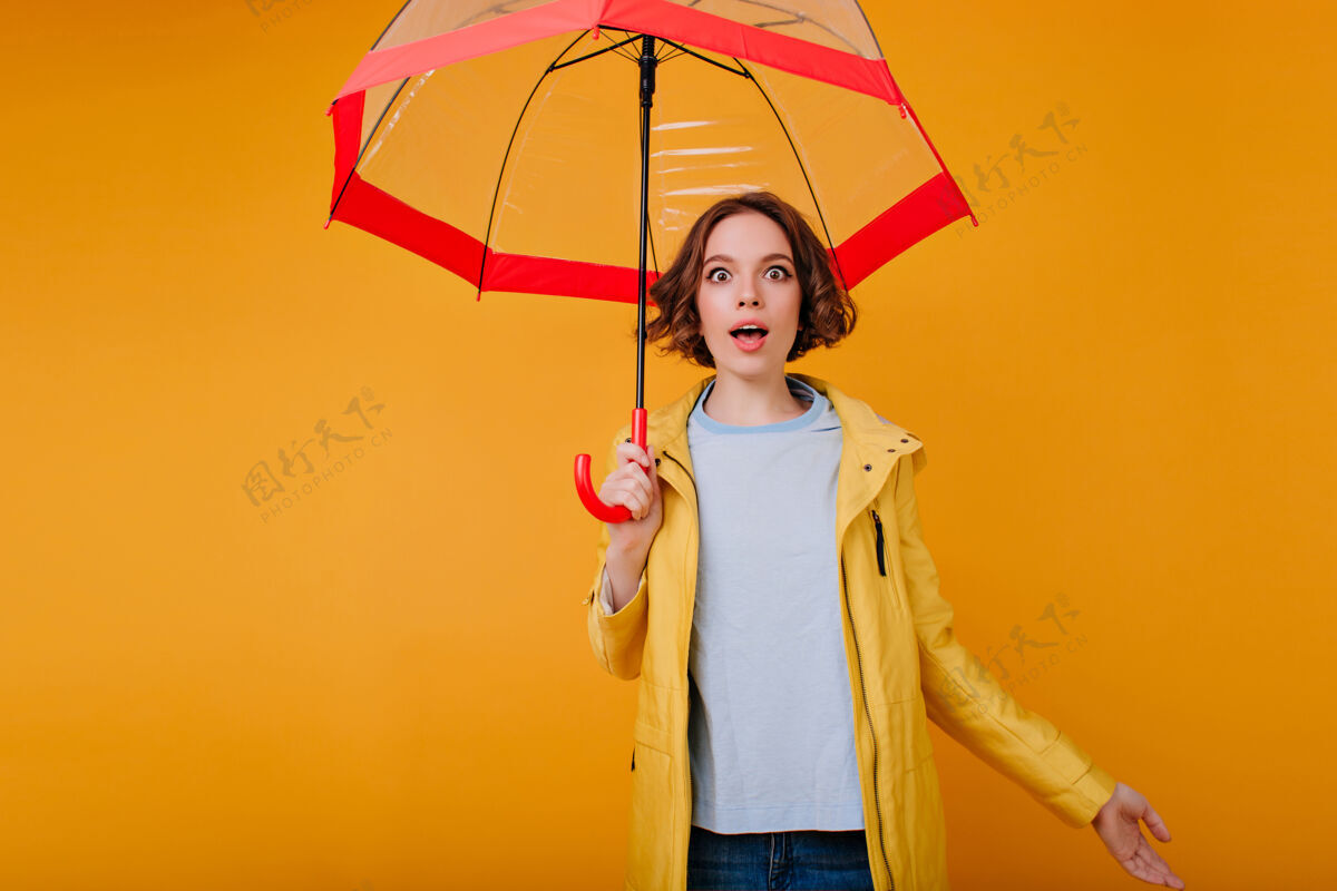 成人身着蓝色衬衫和黄色外套的年轻白种人欣喜若狂 表示惊讶室内照片中 卷曲的梦幻女孩在与阳伞合影时玩得很开心雨姿势惊喜