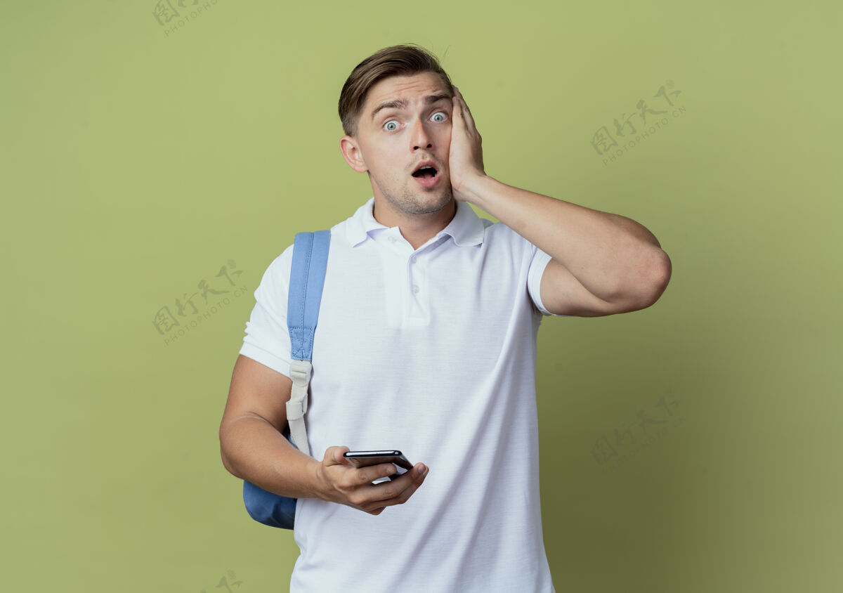 男性吓得年轻帅气的男生背着书包拿着电话 手放在脸颊上孤立的橄榄绿上学生脸颊橄榄